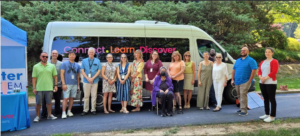 16 smiling people stand in front of a green Westchester Library bus with the words Connect, Learn, Discover on the bus windows.