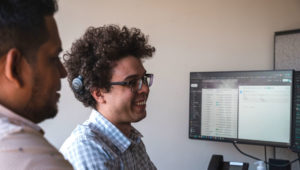 Two adult men smile while looking at a spreadsheet on a computer.